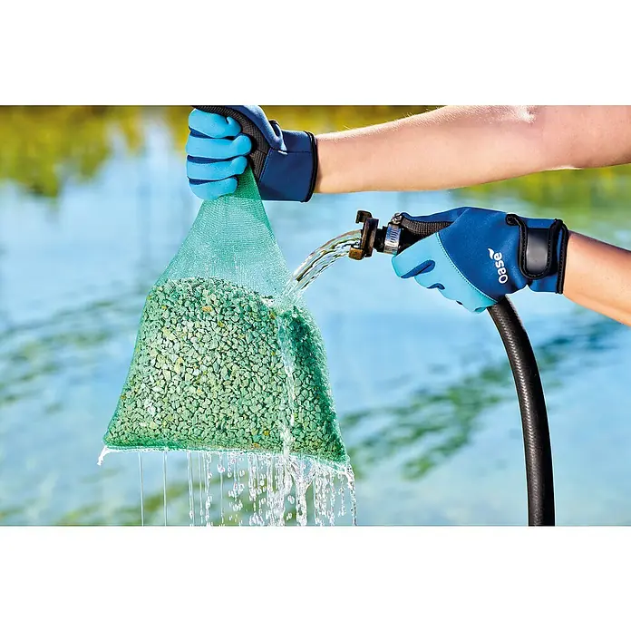 Oase AquaActiv Zeolith Pondlith Filtermaterial - 2,5 kg - Ein Netzbeutel voller Substrat wird mit einem Wasserschlauch gereinigt. Die Oase-Handschuhe schützen die Hände bei der Arbeit.