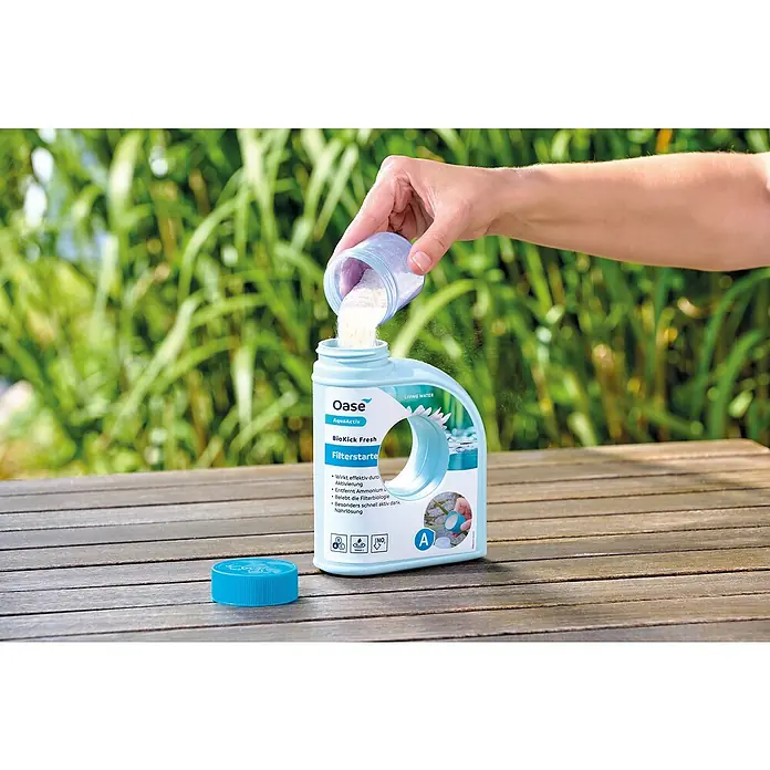 Oase AquaActiv Filterstarter - 500 ml - Eine Hand schüttet Oase BioKick Fresh Filterstarter Pulver in eine blaue Flasche. Das Produkt steht auf einem Holztisch im Freien.