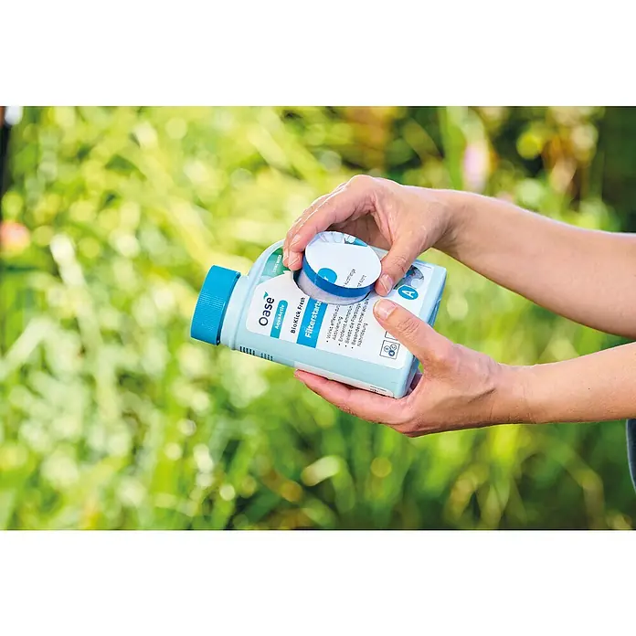 Oase AquaActiv Filterstarter - 500 ml - Hände öffnen eine Flasche Oase BioKick Fresh Filterstarter vor einem verschwommenen grünen Hintergrund.