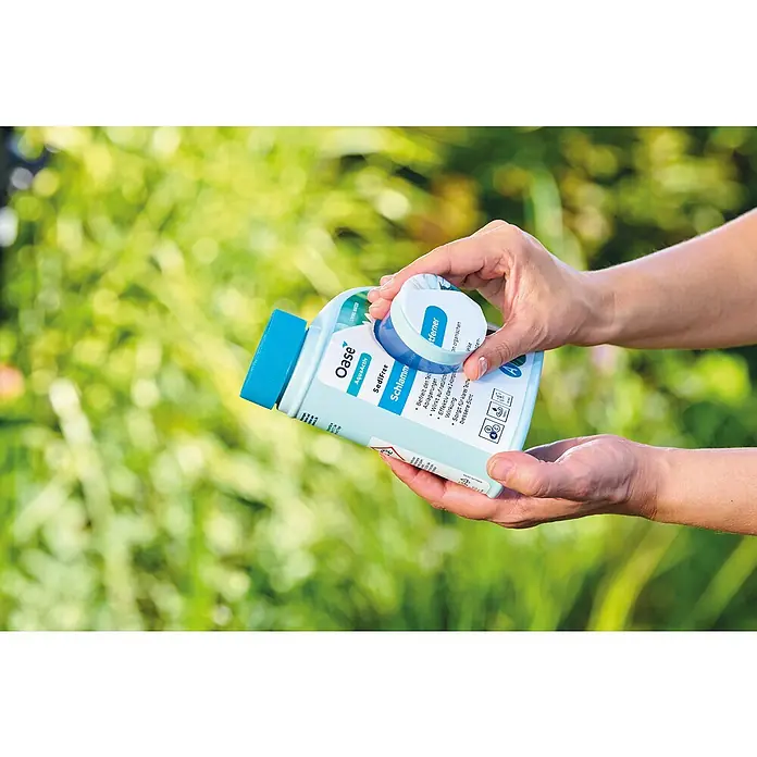 Oase AquaActiv Teichschlammentferner Sedifree - 500 ml - Das Bild zeigt eine Hand, die eine Flasche Oase AquaActiv SediFree Schlammentferner mit geöffnetem Deckel hält, vor einem grünen Hintergrund.