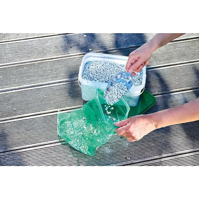 Oase AquaActiv Zeolith Pondlith Filtermaterial - 2,5 kg - Befüllen eines grünen Netzes mit hellgrauem Kies aus einem transparenten Behälter mit einem blauen Messlöffel.