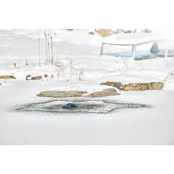 Oase Eisfreihalter IceFree 4 Seasons - Geeignet für: Gartenteiche - Winterlandschaft mit einem solarbetriebenen Springbrunnen in einem teilweise gefrorenen Teich, der Wasser versprüht. Der Springbrunnen ist der zentrale Fokus des Bildes.