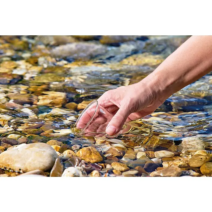 Oase AquaActiv Wasseranalyse-Set - Geeignet für: Gartenteiche - Klares Glas wird mit einer Hand in einen Bach mit Steinen gehalten. Das Wasser umspült das Glas und die Steine am Grund sind sichtbar.