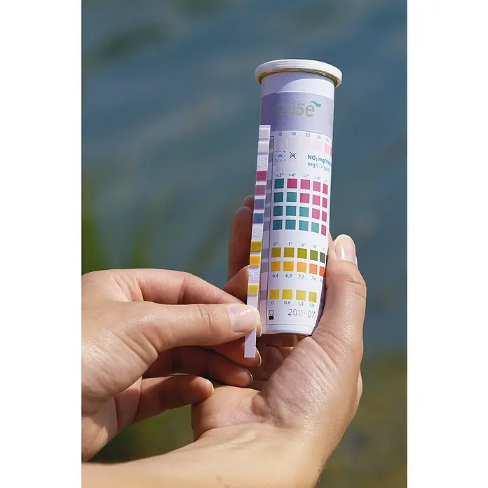 Oase AquaActiv Teststreifen - 50 Stk. - Eine Hand hält einen Wasserteststreifen neben eine Farbskala zur Bestimmung der Wasserqualität.