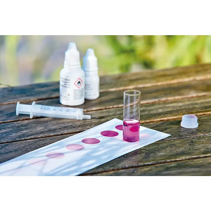 Oase AquaActiv Wasseranalyse-Set - Geeignet für: Gartenteiche - Nahaufnahme eines Wasserqualitätstests mit einer Teströhre mit Flüssigkeit, Flaschen, Spritze und Farbvergleichstabelle auf einem Holztisch.