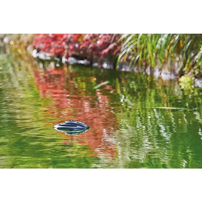 Oase Eisfreihalter IceFree 4 Seasons - Geeignet für: Gartenteiche - Das Bild zeigt einen Teich mit einem schwimmenden, runden Teichroboter, der das Wasser sauber hält. Spiegelungen von Pflanzen sind auf der Wasseroberfläche sichtbar.