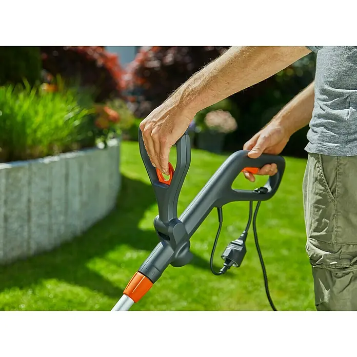 Gardena Elektro-Rasentrimmer PowerCut 650/28 - 650 W, Schnittbreite Faden: 28 cm - Nahaufnahme eines Mannes, der einen Rasentrimmer mit grauem Griff und orangefarbenen Akzenten in einem Garten bedient.