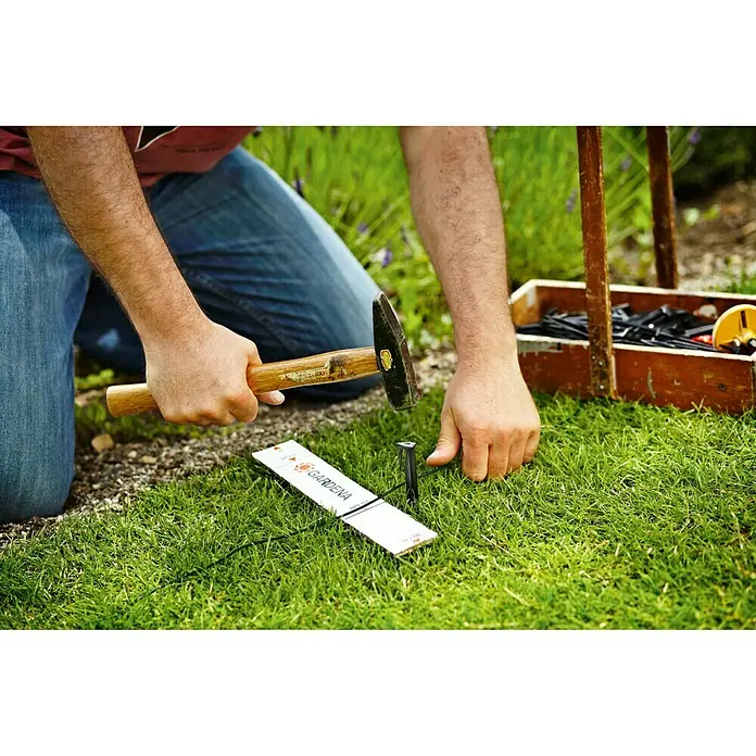 Gardena Begrenzungsdraht für Mähroboter - Länge: 50 m, Gardena Mähroboter - Person nagelt Gardena-Bewässerungsrohrhalter mit Hammer auf Rasen. Praktische Helfer für präzise Gartenbewässerung.