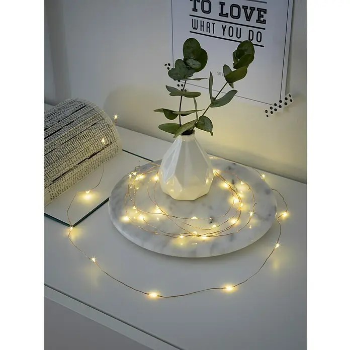 Konstsmide LED-Lichterkette Micro - Innen, Lichtfarbe: Warmweiß, Anzahl Leuchtmittel: 20 Stk., Netzbetrieben - Stimmungsvolle LED-Lichterkette auf einem Nachttisch mit Marmorplatte, einer Vase und Dekoration.