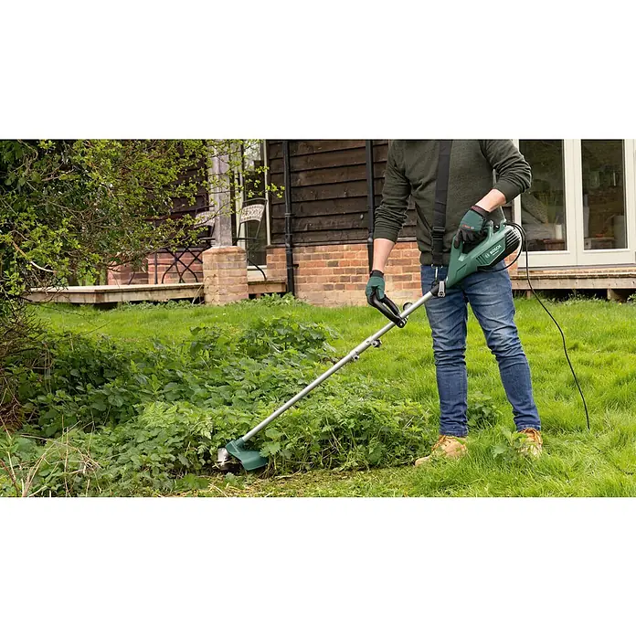 Bosch Elektro-Freischneider AFS 23-37 - 1.000 W, Schnittbreite Faden: 37 cm, Schnittbreite Messer: 23 cm - Ein Mann benutzt einen grünen Bosch Rasentrimmer, um in seinem Garten das Gras zu trimmen. Er trägt Jeans und braune Stiefel.