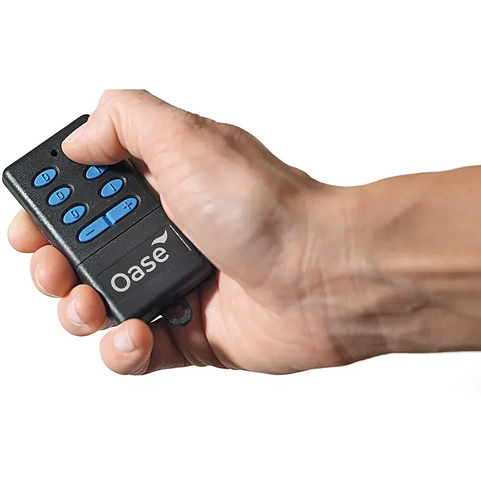 Oase Garten-Steckdose InScenio FM-Master 2 - 3.600 W, 220 V - 240 V - Eine Hand, die eine schwarze Oase-Fernbedienung mit blauen Tasten hält und bedient, fokussiert auf die Bedienung des Geräts.