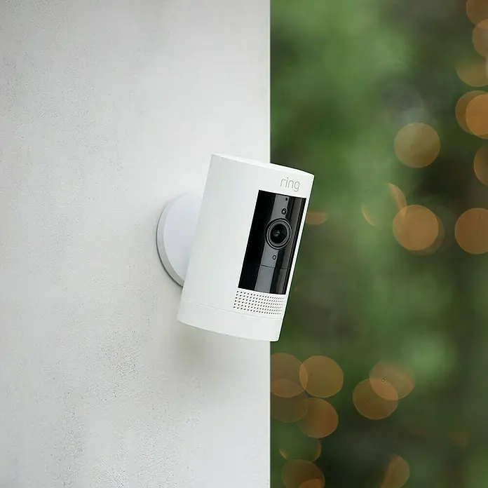 Ring Überwachungskamera außen Stick Up Cam Battery - Akkubetrieben, 1.080 Pixel (Full HD), Weiß - Nahaufnahme einer Ring Sicherheitskamera in Weiß, die an einer Wand befestigt ist. Das Gerät ist kompakt und zylinderförmig, mit einer schwarzen Front.