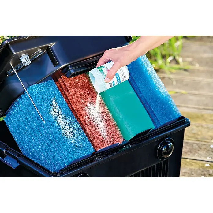 Oase AquaActiv Filterstarter - 100 ml - Ein schwarzer Teichfilter mit verschiedenen Filterschwämmen, in den ein weißes Pulver aus einer Dose gegossen wird.