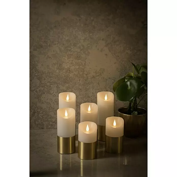 Konstsmide LED-Weihnachtskerzen - Gold, Weiß, Lichtfarbe: Warmweiß, 1 Stk. - Gruppe weißer LED-Kerzen mit goldenen Sockeln, sanft leuchtend auf einer strukturierten Oberfläche neben einer Grünpflanze.