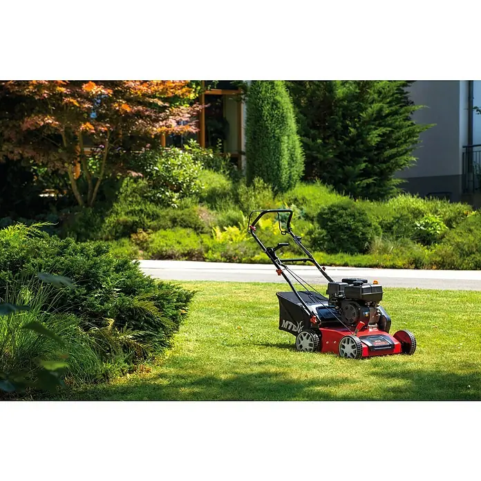 MTD Benzin-Vertikutierer Optima 38 VO - 4 kW, Arbeitsbreite: 38 cm - Ein roter Rasenmäher der Marke MTD steht auf einem gepflegten Rasen vor üppigen Büschen und Bäumen im Garten.