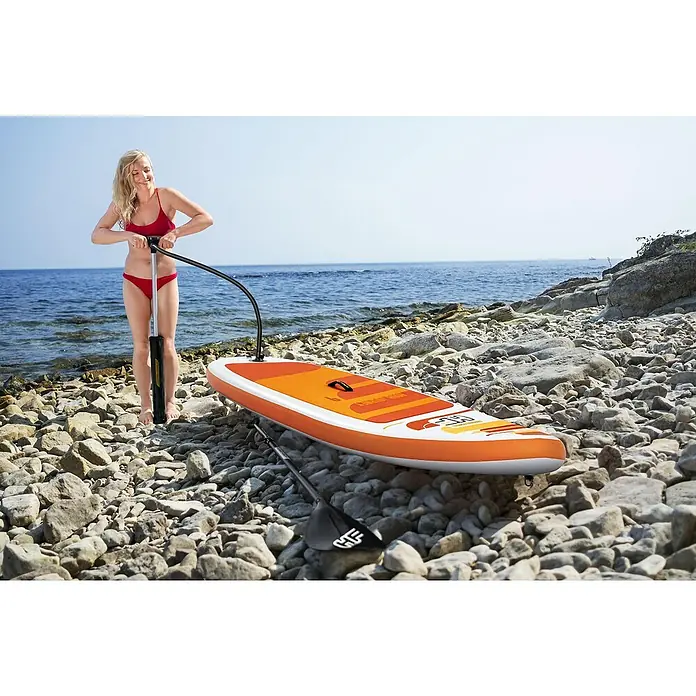 Bestway Hydro Force Juego de paddle surf Aqua Journey - L x An x Al: 274 x 76 x 12 cm, Carga útil: 100 kg, Hinchable, Naranja/Blanco - Una mujer infla una tabla de paddle surf naranja en la orilla. La tabla de paddle surf está en una playa rocosa junto al mar.