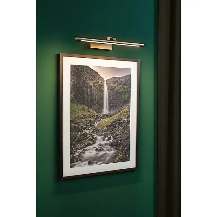 Paulmann LED-Bilderleuchte Mingo - 11 W, L x B x H: 17,5 x 41,5 x 5 cm, Altmessing, Warmweiß - Wandleuchte über einem gerahmten Wasserfallbild an einer grünen Wand. Das Licht beleuchtet das Kunstwerk und betont seine Details.