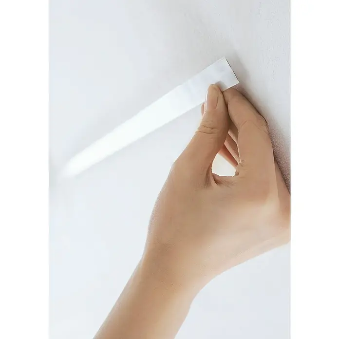 Paulmann Flachkabel - Länge: 400 cm, Geeignet für die Paulmann LED-Bilderleuchten Xana und Adelia, Weiß - Nahaufnahme einer Hand, die ein langes, weißes, rechteckiges LED-Lichtband an einer hellen Oberfläche befestigt.