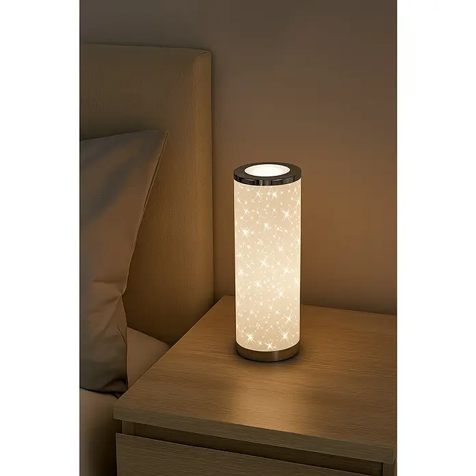 Brilo Lámpara de sobremesa LED redonda Estrellas - 5 W, Ø x Al: 131 x 350 mm, Blanco, Blanco cálido - Esta imagen muestra una lámpara de noche cilíndrica con un estampado de estrellas sobre una mesa de madera junto a una cama.