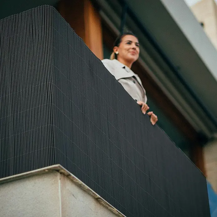 Nortene Sichtschutz LOP - Anthrazit, L x H: 300 x 90 cm - Eine Frau blickt von einem Balkon mit einem blickdichten Sichtschutz aus dunklen Lamellen, der Privatsphäre und modernes Design vereint.
