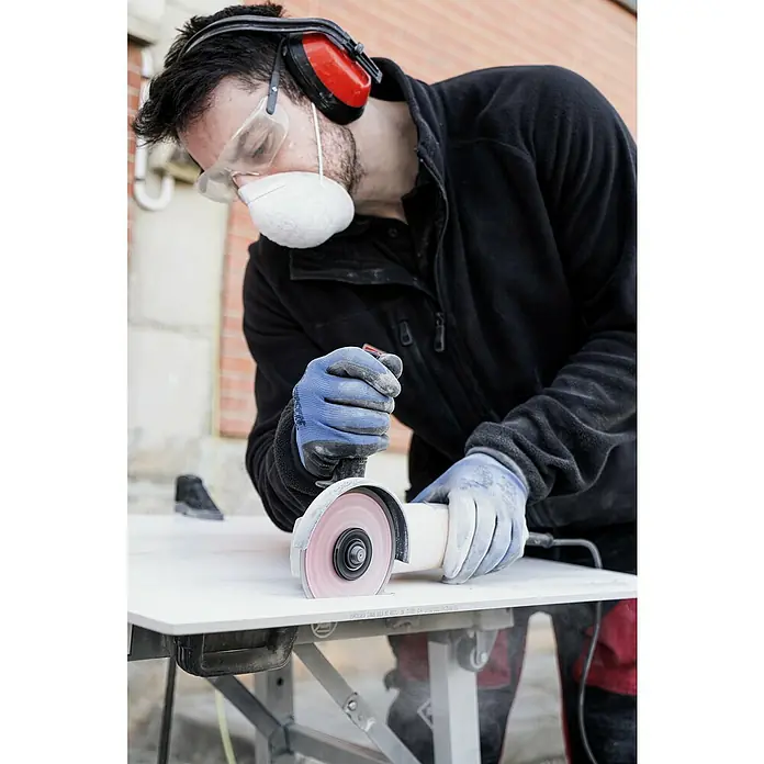 Rubi Disco de corte Slither Superpro - Ø x Es: 125 x 2,2 mm - Un trabajador de la construcción con máscara protectora y protección auditiva utiliza una amoladora para cortar una pieza en una mesa de trabajo.