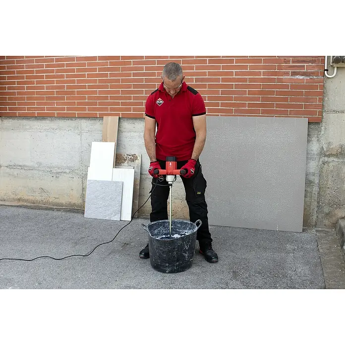 Rubi Mezclador de pintura y mortero Rubimix‑7 MINIMAX - 1.200 W, M 14, Diámetro paleta de mezcla: 120 mm - Hombre mezclando adhesivo para baldosas en un cubo con el mezclador Rubi frente a una pared de ladrillos.