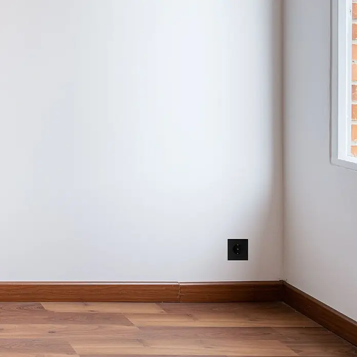 Luceco Oxford Enchufe Schuko pack x3 - Negro, 16 A, 3 ud. - Toma de corriente con carcasa oscura en una pared blanca, complementada con zócalo marrón y suelo de madera.