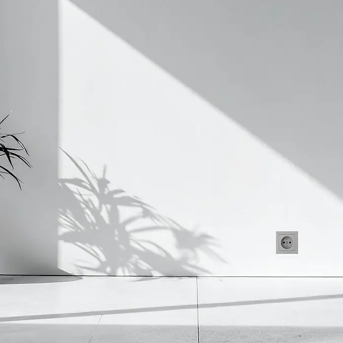Luceco Oxford Enchufe Schuko pack x3 - Gris, 16 A, 3 ud. - Un moderno enchufe gris se integra perfectamente en una pared blanca minimalista, asegurando funcionalidad con una estética elegante.