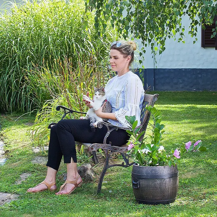 Roto Pflanztopf Bolero - Braun,Schwarz, Außenmaß (Ø x H): 34 x 35 cm, Kunststoff - Eine Frau sitzt auf einer Bank im Garten mit einer dekorativen Pflanzentopf neben sich, der dem Bild eine idyllische Note verleiht.