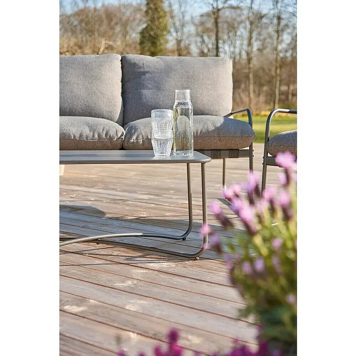 Conjunto de muebles Theodor - 4 pzs., Acero/Poliéster, Antracita - Una mesa de jardín gris con vasos y una botella de agua encima, rodeada de un sofá gris y plantas florecientes.