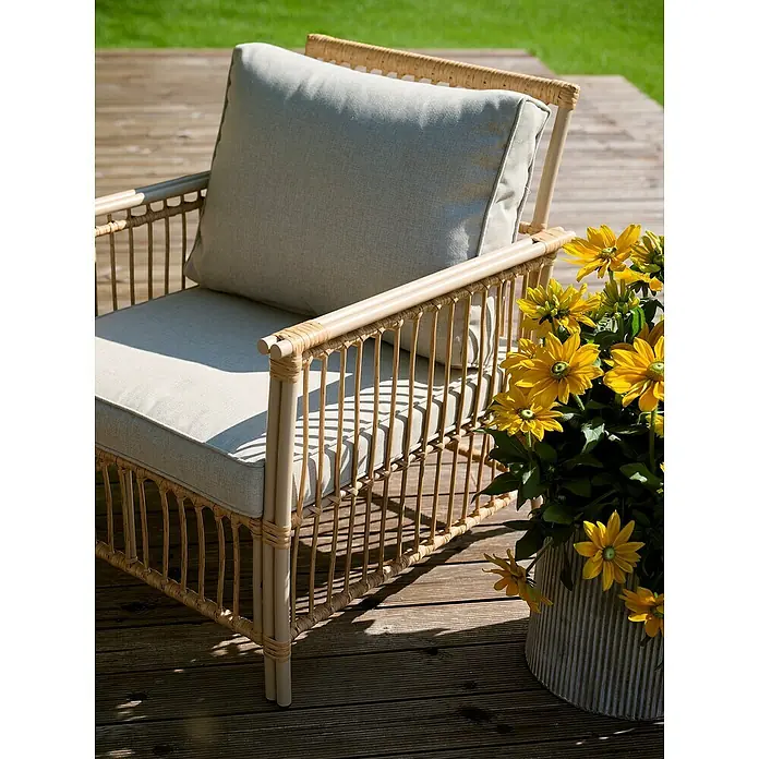 Conjunto de muebles Neel - 4 pzs., Acero/Ratán sintético, Natural/Beige - Primer plano de una silla de mimbre con cojines claros y flores amarillas, sentada en una cubierta de madera.