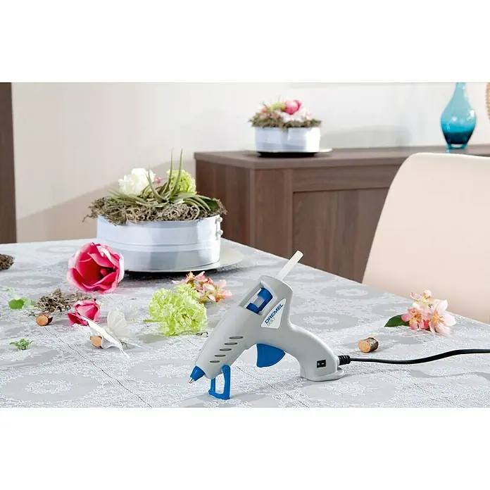 Dremel Pistola termoencoladora Hobby 930-18 - Diámetro cartucho de cola: 7 mm, Tiempo de calentamiento: 5 min - Primer plano de una pistola de pegamento Dremel, ideal para manualidades y proyectos de bricolaje, rodeada de flores y decoraciones en una mesa.
