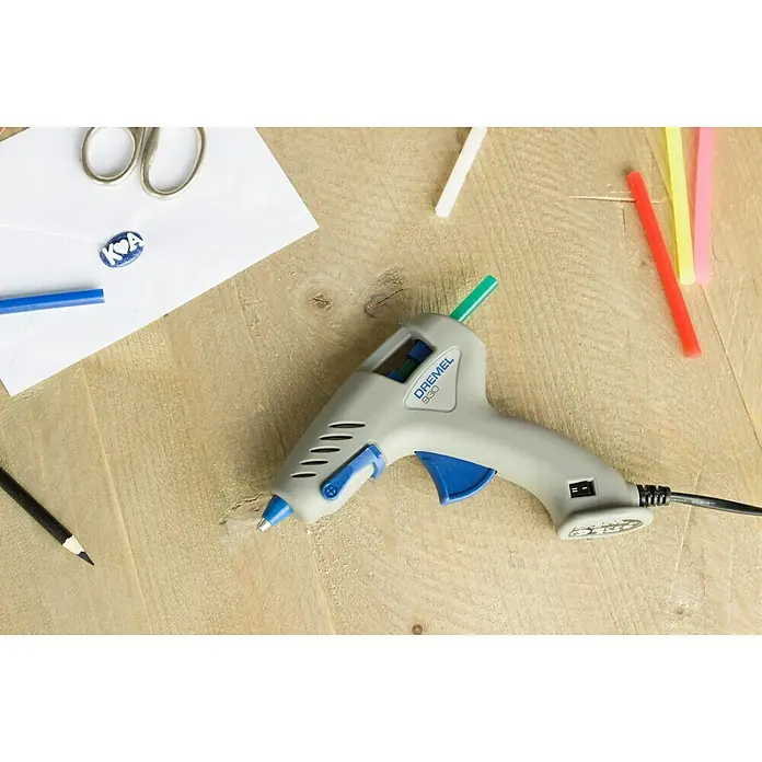 Dremel Pistola termoencoladora Hobby 930-18 - Diámetro cartucho de cola: 7 mm, Tiempo de calentamiento: 5 min - Pistola de pegamento caliente Dremel 930 con detalles azules sobre una mesa de madera con materiales de manualidades.