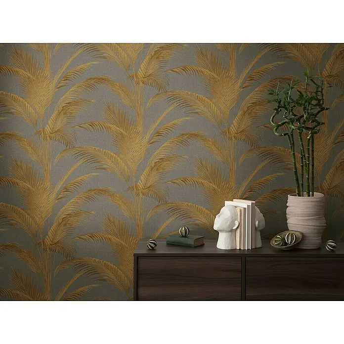 A.S. Création Metropolitan Stories Vliestapete Palmwedel - Grau-Gold-Metallic, Floral, 10,05 x 0,53 m - Nahaufnahme einer Kommode mit einem Deko-Kopf, Büchern, einer Pflanze und kleinen Dekokugeln vor einer Tapete mit Palmenmuster.