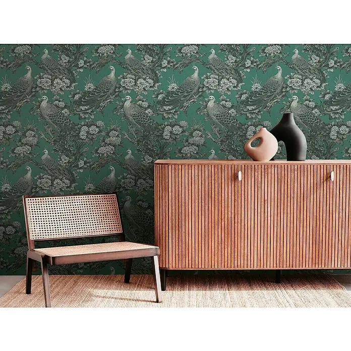 A.S. Création Metropolitan Stories Vliestapete Dschungel - Grau-Grün-Schwarz, Floral, 10,05 x 0,53 m - Interieur mit einem braunen Holzschrank, zwei Vasen und einem Stuhl vor einer grünen Tapete mit Pfauenmuster.