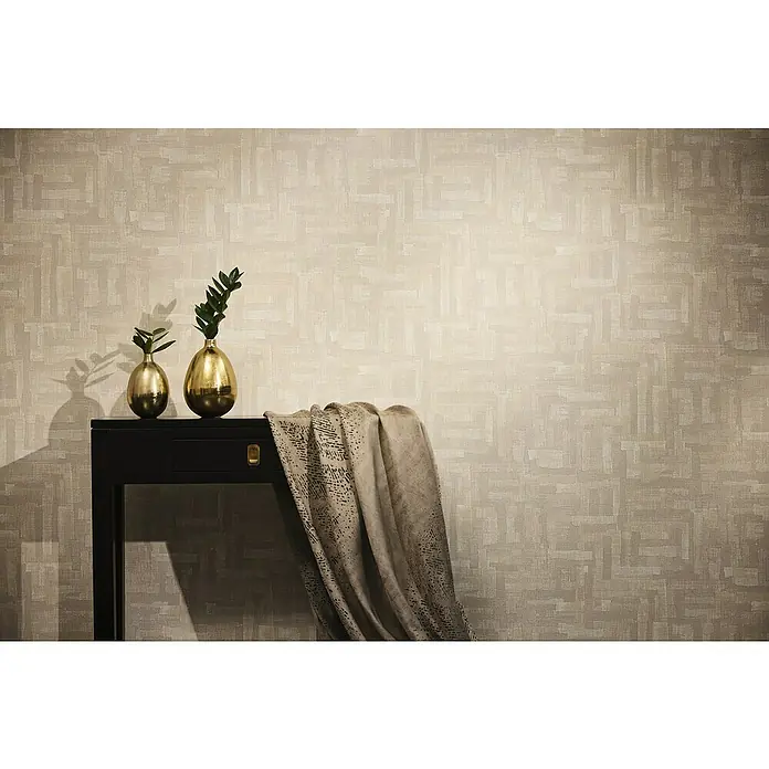 A.S. Création Metropolitan Stories Hot Spots Vliestapete Ethno - Beige-Creme-Grau, Grafisch, 10,05 x 0,53 m - Dekorative Goldvasen mit grünen Zweigen auf einem schwarzen Tisch mit gemusterter Stoffdecke vor beiger Wandtapete.