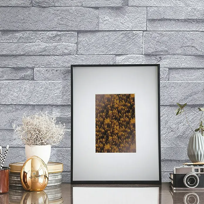 Papel pintado Piedra - Gris, Efecto piedra, 10 x 0,53 m - Foto enmarcada con un motivo de bosque otoñal, rodeada de elementos decorativos sobre una mesa brillante frente a una pared de ladrillo gris.
