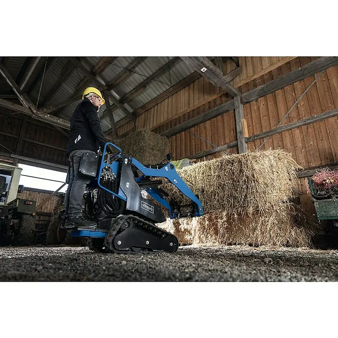 Scheppach Ballenspieß - Passend für: Scheppach Kompaktlader MKL730 - Ein Mann mit Schutzhelm bedient einen Scheppach Mini-Lader vor Heuballen in einer Scheune. Ideal für landwirtschaftliche Arbeiten.