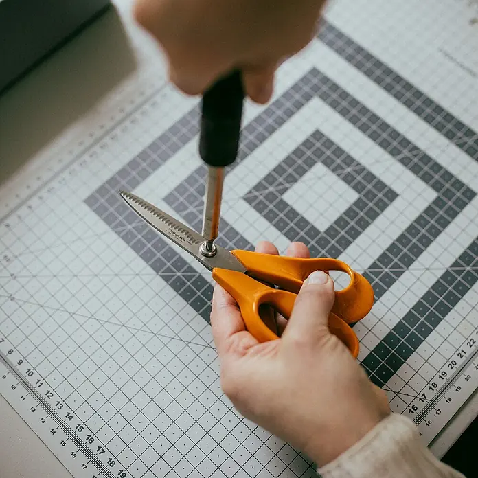 Fiskars Schere Classic - Rechtshänder, Länge: 18 cm, Orange - Detailaufnahme einer Person, die eine orangefarbene Schere mit einem Schraubenzieher auf einer Schneidematte repariert.