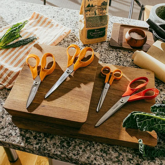 Fiskars Schere Classic - Rechtshänder, Länge: 18 cm, Orange - Verschiedene Fiskars-Scheren auf Holzbrettern mit Lebensmitteln.
