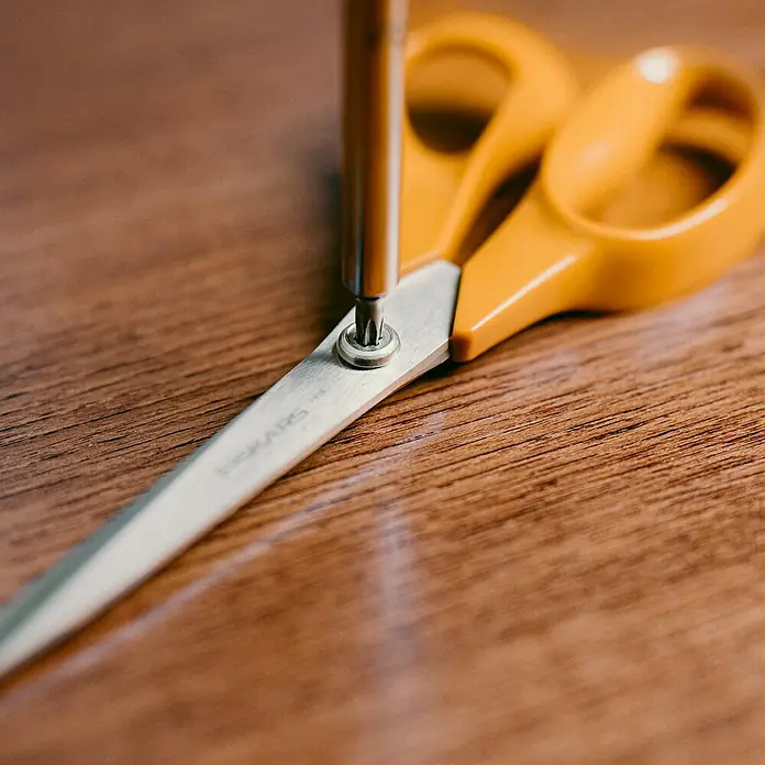 Fiskars Schere Classic - Rechtshänder, Länge: 18 cm, Orange - Nahaufnahme einer Fiskars-Schere, die mit einem Schraubenzieher auf einem Holztisch repariert wird. Ideal für Heimwerker.