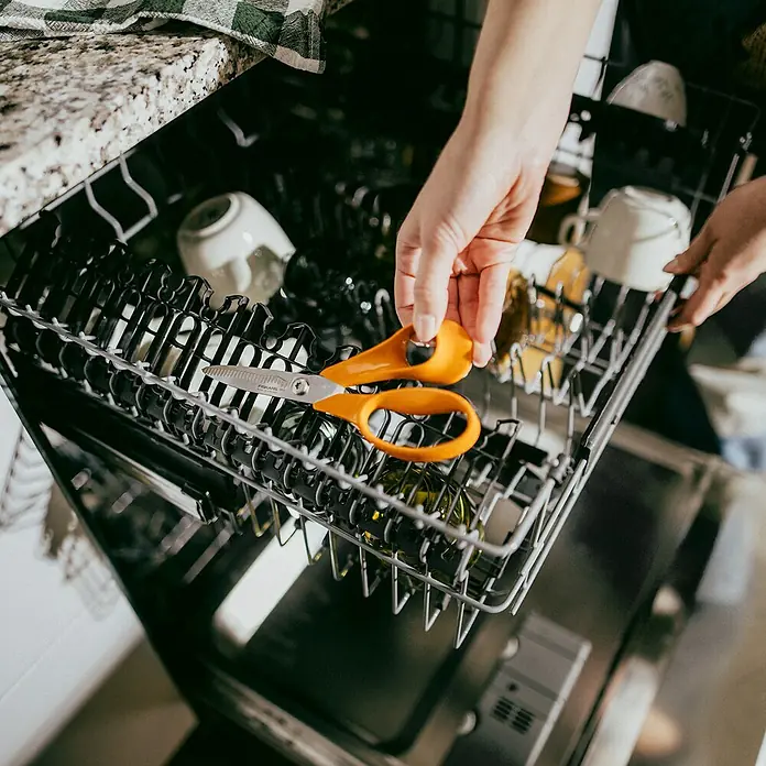 Fiskars Schere Classic - Rechtshänder, Länge: 18 cm, Orange - Eine Person legt eine orangefarbene Fiskars-Schere in eine Geschirrspülmaschine, um sie zu reinigen.