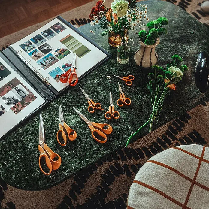 Fiskars Universalschere Classic - Rechtshänder, 25 cm, Farbe: Orange - Anordnung von Scheren verschiedener Größen auf einem grünen Tisch mit Blumen, einem Fotoalbum und einem Hocker, die einen kreativen Arbeitsbereich zeigen.