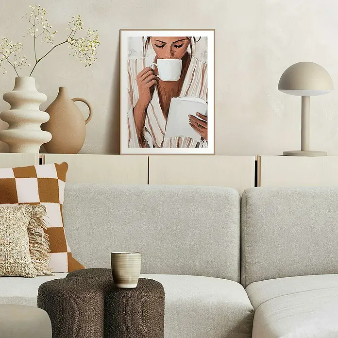 Reinders Bild Slim Frame - Relaxing Time, B x H: 30 x 40 cm - Gemütliches Wohnzimmerambiente mit einem gerahmten Kunstdruck einer Frau, die Kaffee trinkt und liest, über einem beigen Sofa und dekorativen Vasen.