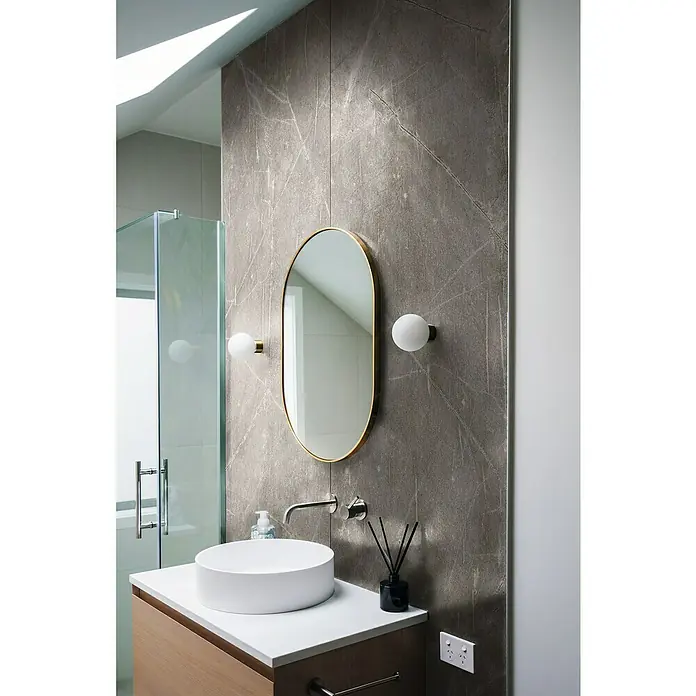 LOGOCLIC Wandpaneel Designwall - Grey Atlantic Marble, 265 x 123 cm - Ein stilvolles Badezimmer mit einem ovalen Spiegel mit goldenem Rahmen, der zwischen zwei Kugelwandleuchten hängt, über einem modernen Aufsatzwaschbecken.