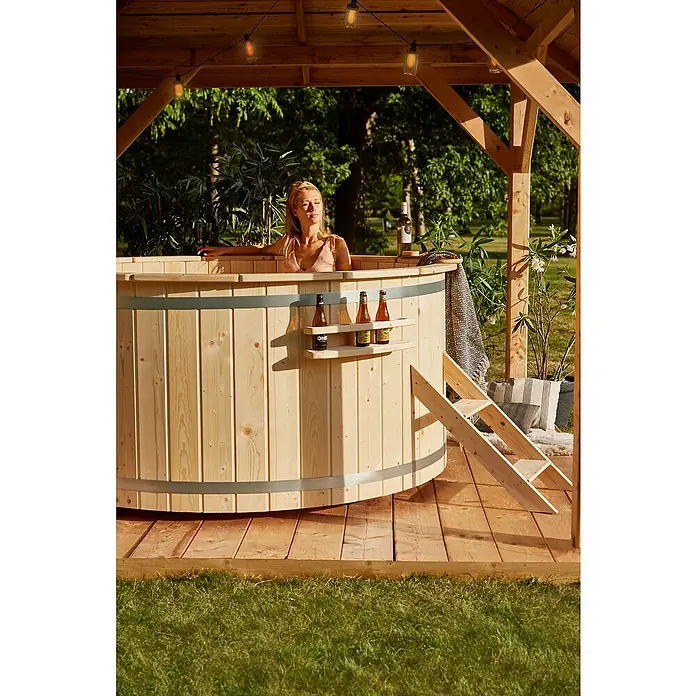Tuindeco Hot Tub - 200 cm, Fichte, Natur, Max. Personenzahl: 6 Stk. - Ein hölzerner Whirlpool im Freien mit einer Frau, Getränken und dekorativer Beleuchtung für ein entspannendes Erlebnis.