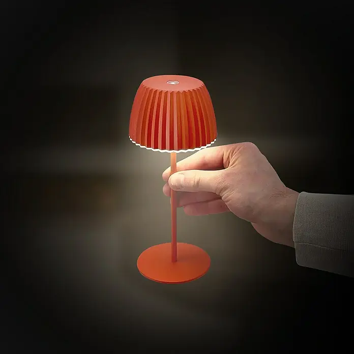 Brilo Riffle Akku-Tischleuchte - Ø x H: 8,5 x 20,1 cm, Orange, Warmweiß, 1,5 W, IP44, Mini - Eine orangefarbene Tischlampe mit plissiertem Schirm wird von einer Hand gehalten und spendet warmes Licht.
