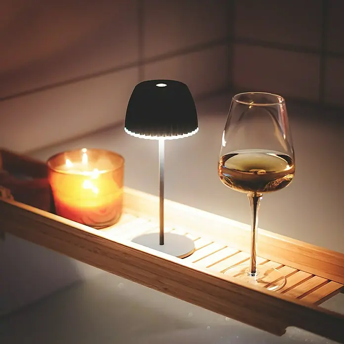 Brilo Riffle Akku-Tischleuchte - Ø x H: 8,5 x 20,1 cm, Anthrazit, Warmweiß, 1,5 W, IP44, Mini - Ein stimmungsvolles Bild mit einer Lampe, einer Kerze und einem Glas Wein auf einem Badewannenbrett. Die sanfte Beleuchtung erzeugt eine entspannende Atmosphäre.
