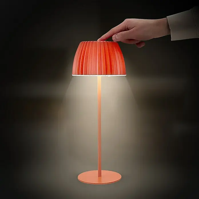 Brilo Riffle Akku-Tischleuchte - Ø x H: 14,3 x 36,4 cm, Orange, Warmweiß, 2 W, IP44, Classic - Eine moderne orangefarbene Tischlampe mit plissiertem Lampenschirm wird von einer Hand berührt, um sie einzuschalten.