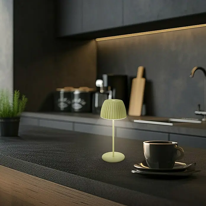 Brilo Riffle Akku-Tischleuchte - Ø x H: 8,5 x 20,1 cm, Grün, Warmweiß, 1,5 W, IP44, Mini - Grüne Tischlampe mit plissiertem Lampenschirm auf einer Küchentheke neben einer Kaffeetasse.